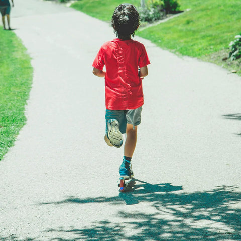 Child riding a kick scooter on a sunny park path; red shirt and blue shorts.