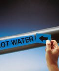 Hand applying a Top Shelf Self Stick Compressed Air Pipe Marker 1X8 Inch White On Blue to hot water piping.