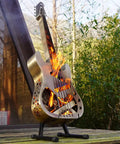 Guitar-shaped stainless steel fire pit with flames, standing on a wooden platform outdoors.