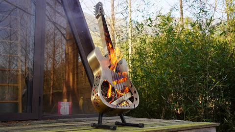 Guitar-shaped stainless steel fire pit with flames, standing on a wooden platform outdoors.