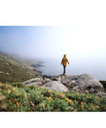 Person in yellow jacket stands on rocky cliff overlooking foggy ocean, demonstrating the rugged weather-sealed camera's outdoor use.