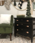 Black dresser with 3 Inch Bin Cup Drawer Cabinet Pulls In Antique Copper Finish beside green chair, holiday decor on wall.