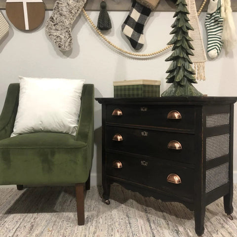 Black dresser with 3 Inch Bin Cup Drawer Cabinet Pulls In Antique Copper Finish beside green chair, holiday decor on wall.