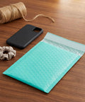 Teal 5x6 Inch Poly Bubble Mailers With Self Seal Adhesive Waterproof Padded Shipping Bags For Small Biz Packing Durable Bubble Envelopes For Secure Item Protection 100 Count shown on wooden table with accessories.