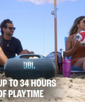 Two people sitting on a beach with a black Hardest Hitting Portable Waterproof Speaker With Pro Sound And AI Boost between them.
