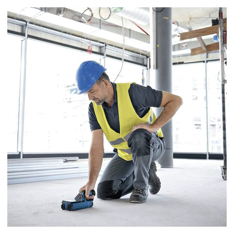 Man using Hardcore Stud Finder Wall Scanner on floor in construction site, wearing safety vest and helmet, focused on accuracy.