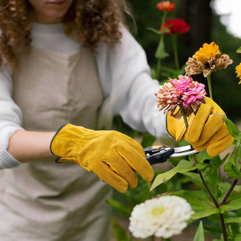 Person wearing Tough Cowhide Leather Work Gloves With Flex Grip For Wood Cutting, Construction, Driving, And Gardening For Men And Women, Breathable Sweat Wicking Palm, Durable Outdoor Gloves, Large Gold 1 Pair, trimming flowers in garden.