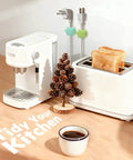 White coffee maker and toaster on wooden counter with two slices of toast, coffee cup, and decorative pinecone tree.