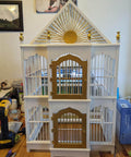 White and gold birdcage with sunburst design and arched doors on a wooden table surrounded by tools and objects.