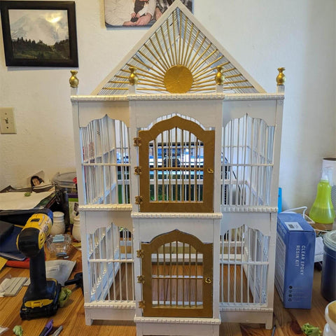 White and gold birdcage with sunburst design and arched doors on a wooden table surrounded by tools and objects.