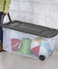 Rugged Pull Along 160 Quart Rolling Storage Tote With Transparent Bottom, holding colorful beach toys, on wheels.