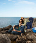 Couple sitting on rocks by the sea with a heavy duty directors chair, side table, pockets, and cooler in the background.
