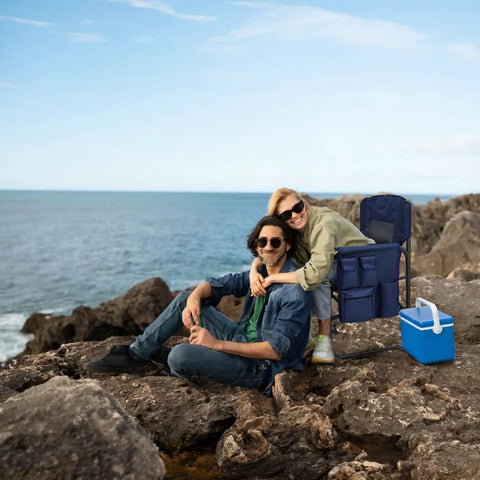 Couple sitting on rocks by the sea with a heavy duty directors chair, side table, pockets, and cooler in the background.