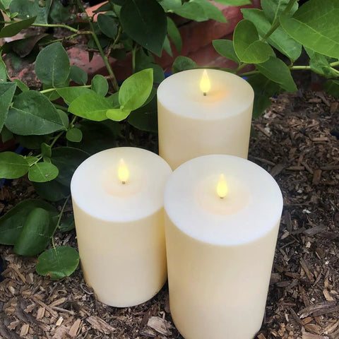 Waterproof Outdoor Flameless Pillar Candles 3 Pack In Ivory Resin, 3x4, 3x5, And 3x6 Inches, arranged on mulch with LED flicker