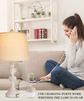 Coastal Farmhouse Table Lamp with USB port, washed white finish, beige shade, on a living room table. Woman using laptop and phone.