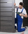 Person in blue overalls repairing a dark wooden door with a green power drill inside a room.