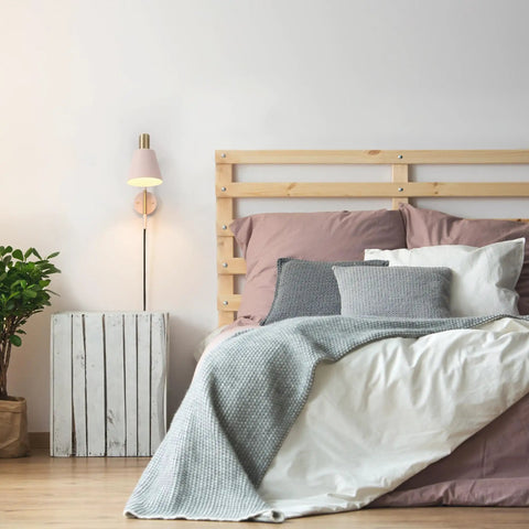 Cleo 1 Light Wall Sconce with Plug-In or Hardwire Setup, blush pink and matte brass accents, mounted by a wooden bed in styled bedroom.