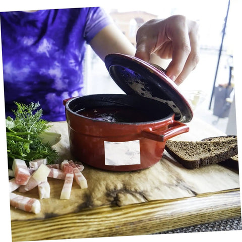 Person opening a red ceramic pot with cooked dish inside, surrounded by herbs, bread, and sliced bacon on a wooden board.