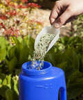 Hand pouring seeds into Heavy Duty Hand Held Spreader With Adjustable Hole Size For Seeds Fertilizer Salt Ice Melt And Pest Control Granules 2 Liter Capacity Includes Scoop Durable Garden Tool For Lawn Garden And Driveway Maintenance Blue