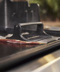 Heavy Duty Weber Griddle Press For Perfect Bacon, Smash Burgers And Crispy Paninis over bacon on a hot griddle.