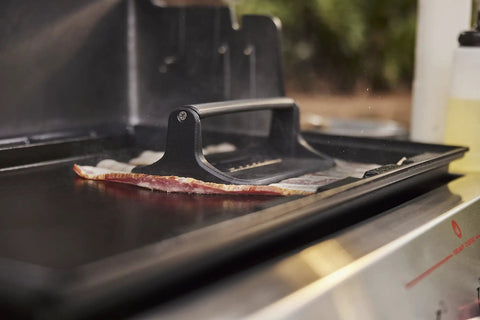 Heavy Duty Weber Griddle Press For Perfect Bacon, Smash Burgers And Crispy Paninis over bacon on a hot griddle.