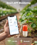 A hand holding a smartphone displaying an app, next to the 2-In-1 Wireless Soil Meter With WiFi And Bluetooth in a garden bed.