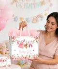 13 Inch Pink Floral Baby Gift Bag With Sturdy Handles Includes Matching Card And Three Sheets Of Tissue Paper The Perfect Gift Bag For Girl Baby Showers And Kiddo Birthday Parties held by a woman in a nursery setting.