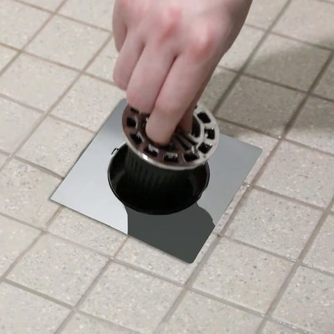 Hand inserting a plastic catch net into a square chrome shower drain, designed to trap hair and prevent clogs.