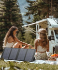 Two women sitting on a picnic blanket next to a 40W Foldable Solar Panel Charger For Power Stations And Camping Gear, RV in background.