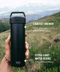Hand holding Ultralight Travel French Press Coffee Maker For On The Go Brews, Vacuum Insulated Stainless Steel 16 Ounce Capacity, Matte Meteorite Black, Ideal For Camping, Commuting, Or Office Breaks, in a natural outdoor setting.