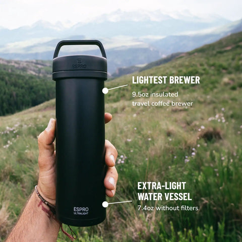 Hand holding Ultralight Travel French Press Coffee Maker For On The Go Brews, Vacuum Insulated Stainless Steel 16 Ounce Capacity, Matte Meteorite Black, Ideal For Camping, Commuting, Or Office Breaks, in a natural outdoor setting.