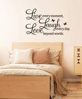 Bedroom view with 'Live Every Moment Laugh Every Day Love Beyond Words' wall sticker above bed, wooden decor accents.