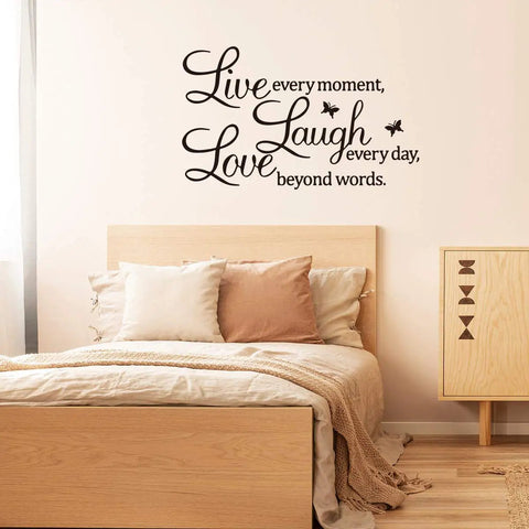 Bedroom view with 'Live Every Moment Laugh Every Day Love Beyond Words' wall sticker above bed, wooden decor accents.