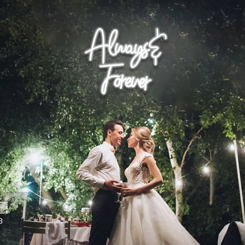 Wedding couple dancing under 'Always & Forever' neon sign in outdoor setting with twinkling lights and lush greenery background.