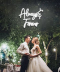 Couple dancing under 'Always & Forever' neon sign, outdoor wedding setting at night, surrounded by trees and lights.