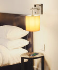 Bedside Wall Sconce With USB Port And AC Outlet, White Fabric Shade, Hardwired Mount Light, shown in bedroom setup, bulb lit