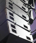 Stacked black speaker boxes with cables on a stage, showing front panels and mounting brackets in a live event setting.