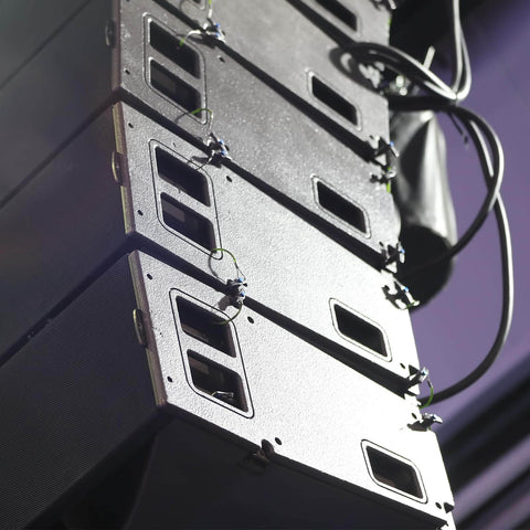Stacked black speaker boxes with cables on a stage, showing front panels and mounting brackets in a live event setting.