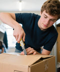 Young man using box cutter to open package in dorm room setting.