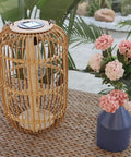 Large Outdoor Solar Lantern With Rattan And Bamboo Build Dope Glow For Garden Patio And Yard Waterproof Hanging Lantern With Handle And Edison Bulb Openwork Design For Tabletop Decoration on woven table next to flowers.