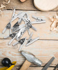 Super Heavy Duty 12-Pc Metal Spring Clamps Kit With Tough White Tips Perfect For Woodworking, DIY Panels, And Backdrop Stands To Hold Projects Steady In Home Shop Or Studio Setting, 12-piece metal clamps on wood table