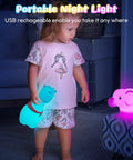 Child holding a glowing blue animal-shaped lamp, standing beside a pink elephant lamp on a stool in a dimly lit room.