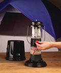 Person adjusting QuickPack Deluxe 1000 Lumen Propane Lantern With Carry Case on a wooden table next to a blue tent.
