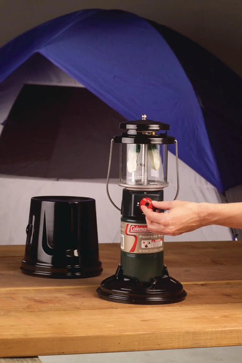 Person adjusting QuickPack Deluxe 1000 Lumen Propane Lantern With Carry Case on a wooden table next to a blue tent.