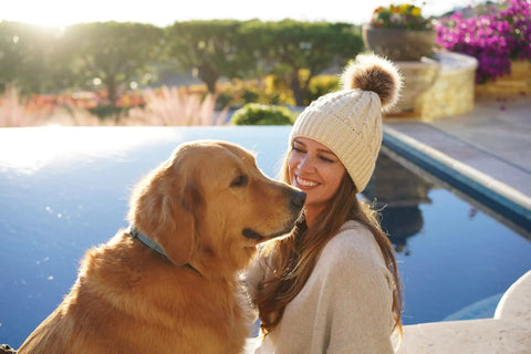 Woman wearing Cozy Cable Knit Beanie Hats for Women With Faux Fur Pom Poms 2 Pack Winter Set in Gray and Cream beside a pool with a dog