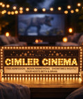 Outdoor home cinema setup with illuminated 'Cimler Cinema' welcome sign, popcorn, and cozy seating under string lights.