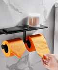 Double Roll Toilet Paper Holder With Shelf Matte Black Brass Finish, holding orange rolls, mounted on marble wall, with phone and jar on shelf.