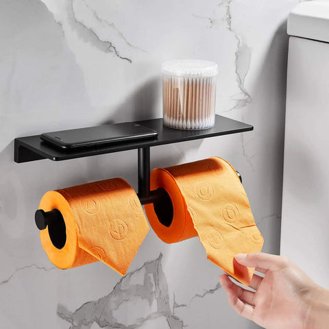 Double Roll Toilet Paper Holder With Shelf Matte Black Brass Finish, holding orange rolls, mounted on marble wall, with phone and jar on shelf.