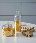Front view of two clear glass bottles with gold leaf on lower halves; wooden bead garland on white table.