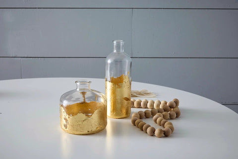 Front view of two clear glass bottles with gold leaf on lower halves; wooden bead garland on white table.